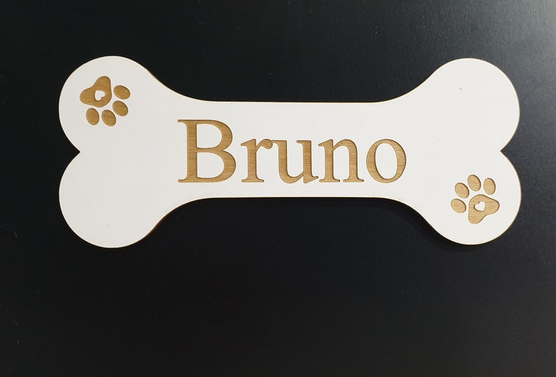 May include: White wooden dog bone with the name "Bruno" engraved in gold lettering. The bone has two gold paw prints on either side of the name.