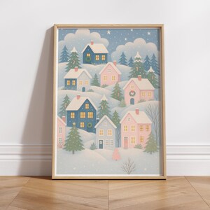 May include: A framed print featuring a winter village scene. The artwork depicts several houses in shades of blue and pink, nestled among snow-covered hills and evergreen trees. The sky is a light blue with falling snow and fluffy clouds.