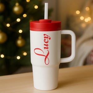 K&ouml;nnte beinhalten: Wei&szlig;er Reisebecher mit rotem Deckel und Griff, mit dem Namen "Lucy" in roter Schrift. Enth&auml;lt einen wei&szlig;en Strohhalm. Der Becher ist f&uuml;r kalte Getr&auml;nke konzipiert und ideal f&uuml;r unterwegs.