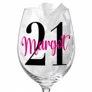 May include: A clear wine glass with the number "21" in black and the name "Margot" in pink script. The glass is filled with white tissue paper. Ideal for a birthday celebration.