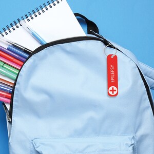 May include: A light blue backpack with a red medical alert tag that says "EPILEPSY" with a white cross on it. The backpack has a zipper and a pocket on the front. The backpack is filled with colorful pens and a notebook.