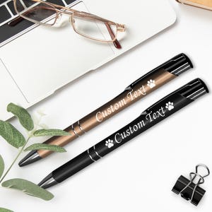 May include: Two personalized pens with a paw print design. One pen is black with a silver clip and the other is gold with a silver clip. Both pens have the text "Custom Text" engraved on them.