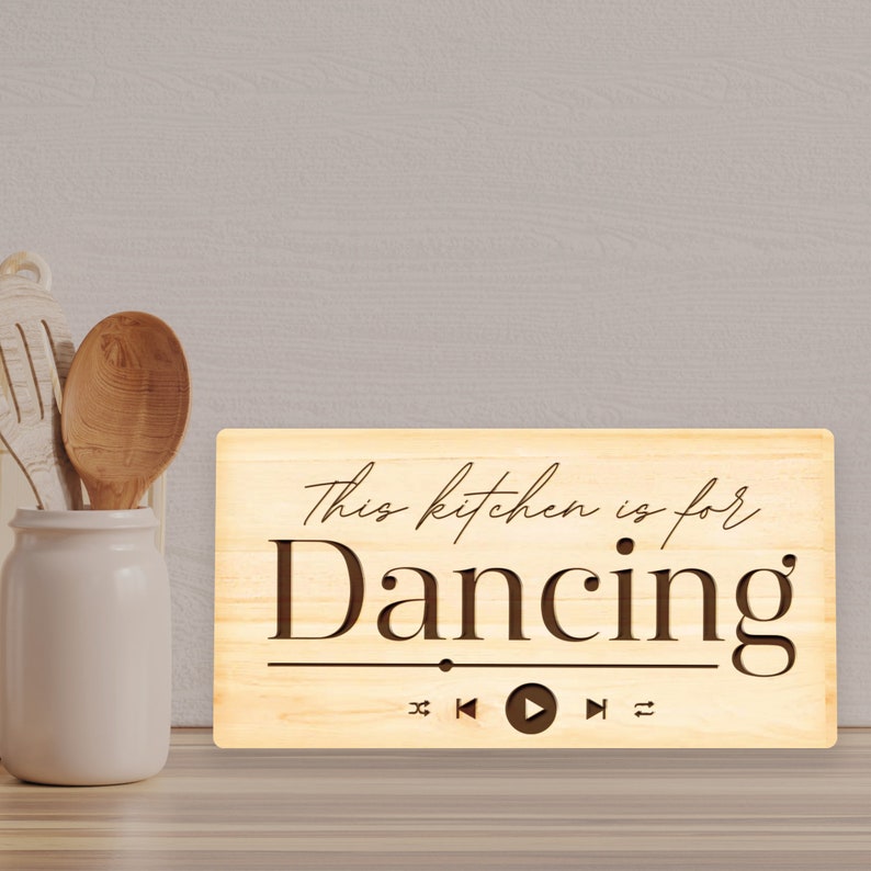 May include: A light-colored wooden sign with the text "This kitchen is for Dancing" in a decorative font. Below the text are music player control icons. A white ceramic jar with wooden spoons is to the left.