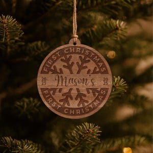 May include: A round, wooden Christmas ornament with the text "First Christmas" and "Mason's" engraved on it. The ornament features a snowflake design and the year "2025". It hangs from a natural twine string.
