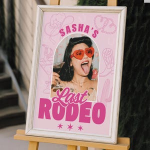 May include: A framed pink sign with the text "Sasha's Last Rodeo" in bold pink letters. The sign features a photo of a person wearing heart-shaped sunglasses and holding a drink. Cowboy-themed illustrations and stars complete the design.