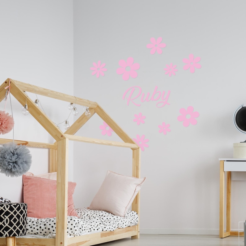 Puede incluir: Calcoman&iacute;as de pared de flores rosas con el nombre "Ruby" en escritura cursiva. Las calcoman&iacute;as est&aacute;n en una pared blanca sobre un marco de cama de madera.