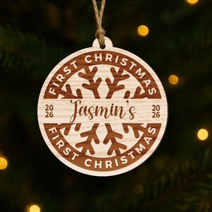 May include: A wooden Christmas ornament with the text "First Christmas" and "Jasmin's" engraved in a circular design. The ornament has a snowflake design in the center and the year 2026.