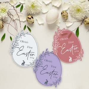 May include: Three pastel-colored oval-shaped Easter decorations with a floral wreath design. Each decoration has the text "Olivia's First Easter 2024" with a bunny silhouette.