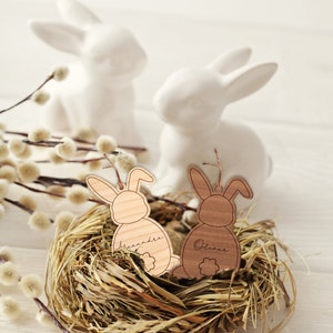 May include: Two wooden bunny ornaments with the names "Alexandre" and "Oliver" engraved on them, nestled in a bird's nest made of straw.