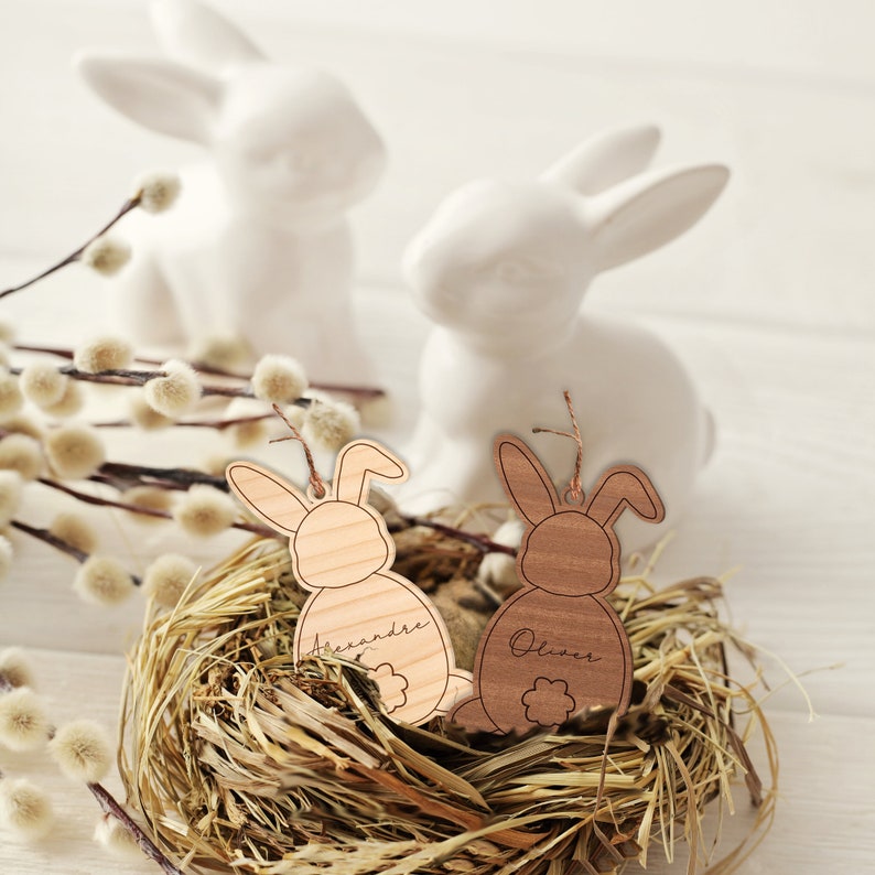 May include: Two wooden bunny ornaments with the names "Alexandre" and "Oliver" engraved on them, nestled in a bird's nest made of straw.
