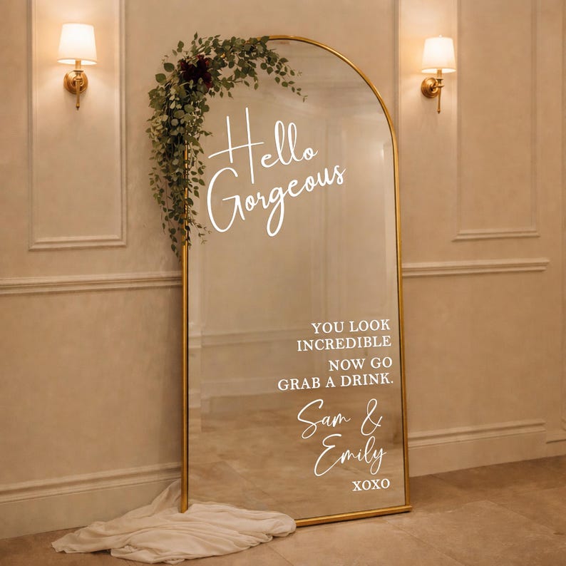 May include: A large, arched mirror with a gold frame. The mirror has the words "Hello Gorgeous" and other text in white script. Greenery and burgundy flowers adorn the top. A white fabric rests at the base of the mirror.