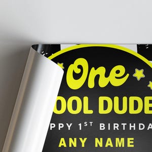 May include: A black and yellow birthday sign with the words "One Cool Dude" in yellow. The sign also includes the text "Happy 1st Birthday" and space for a name. The sign is partially rolled up, revealing a white edge.