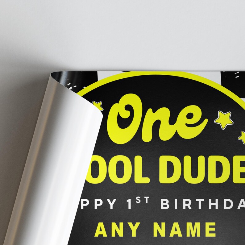 May include: A black and yellow birthday sign with the words "One Cool Dude" in yellow. The sign also includes the text "Happy 1st Birthday" and space for a name. The sign is partially rolled up, revealing a white edge.