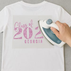 May include: White t-shirt with a pink glitter design that reads "Class of 2022 GEORGIA." An iron is being used to apply the design. The shirt is made of a soft, comfortable material.