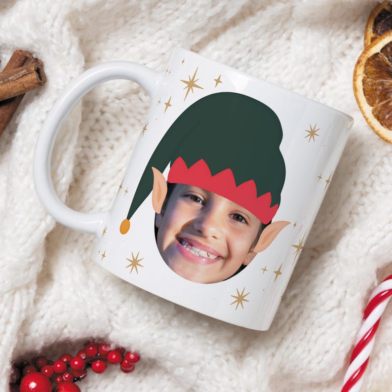 May include: White ceramic mug featuring a child's face wearing an elf hat with a green cap and red trim. The mug is decorated with gold starbursts, ideal for the festive season.