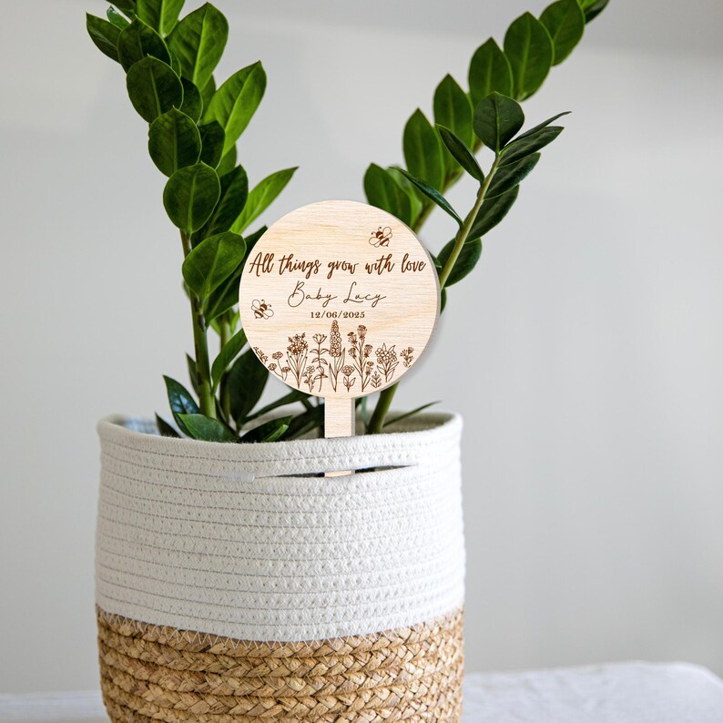 K&ouml;nnte beinhalten: Ein Pflanzenstab aus Holz mit einem runden, gravierten Schild, auf dem "All things grow with love Baby Lacy 12/06/2023" steht. Das Schild zeigt ein florales Design mit Bienen und ist an einem Holzstab befestigt.