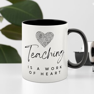 Puede incluir: Taza de cer&aacute;mica blanca con interior y asa negros. La taza presenta el texto "Teaching is a work of heart" en fuente cursiva, junto con un dise&ntilde;o de coraz&oacute;n de huellas dactilares. La taza es de tama&ntilde;o est&aacute;ndar para bebidas calientes.