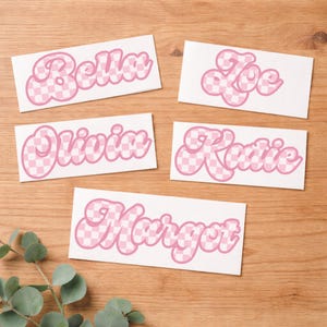 May include: Five white rectangular stickers with names in a retro font. The names are Bella, Zoe, Olivia, Katie, and Margot. Each name is in a pink, checkered pattern with a pink outline.
