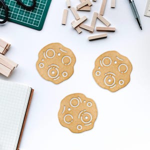 Puede incluir: Tres posavasos de madera con un dise&ntilde;o circular que presenta un patr&oacute;n de c&iacute;rculos y l&iacute;neas. Los posavasos son de color marr&oacute;n claro y tienen un acabado de madera natural.