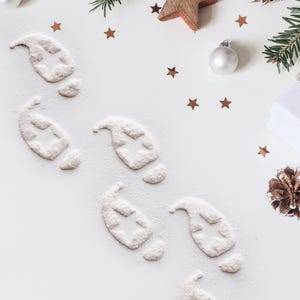 May include: Overhead shot of a white surface decorated with Christmas elements. Footprints made of powder with star cutouts are scattered across the surface. Additional decorations include star-shaped confetti, a wooden star, pine needles, and a silver ornament.
