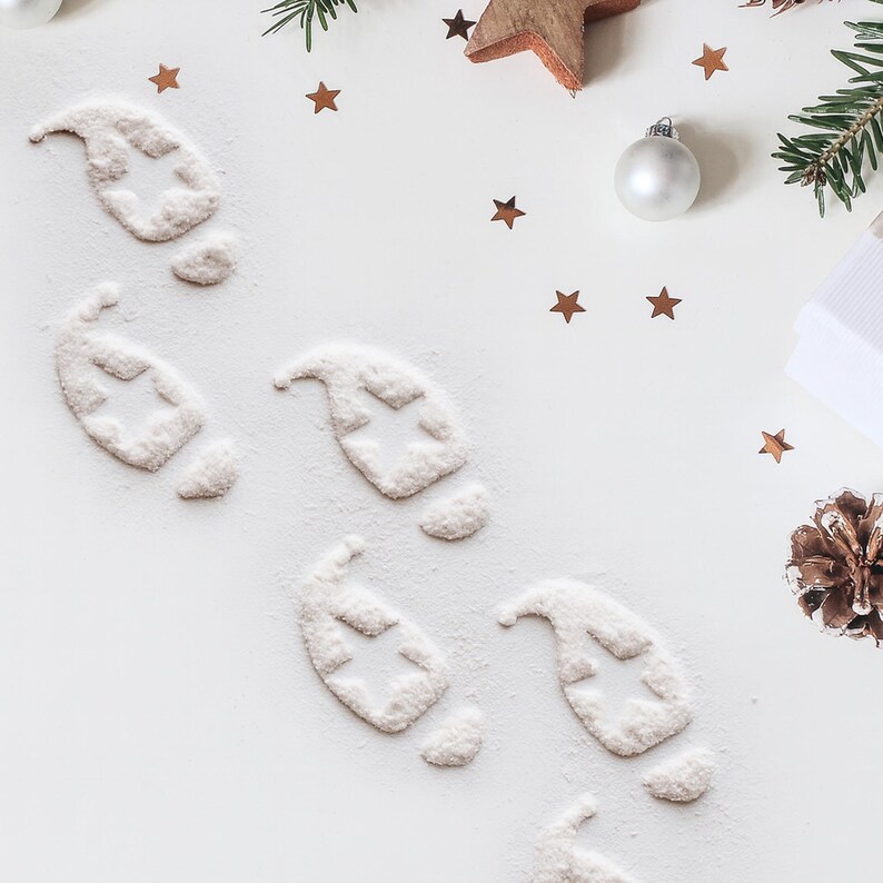 May include: Overhead shot of a white surface decorated with Christmas elements. Footprints made of powder with star cutouts are scattered across the surface. Additional decorations include star-shaped confetti, a wooden star, pine needles, and a silver ornament.