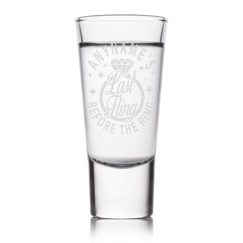 May include: Clear shot glass with etched text reading "ANYNAME'S Last Fling BEFORE THE RING" around a diamond ring graphic. The glass is cylindrical with a thick base and is filled with a clear liquid. Ideal for a hen party or wedding celebration.