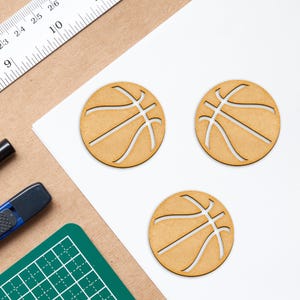 Puede incluir: Tres recortes de madera de balones de baloncesto sobre una superficie blanca. Los balones de baloncesto son marrones y tienen un contorno blanco de las costuras.