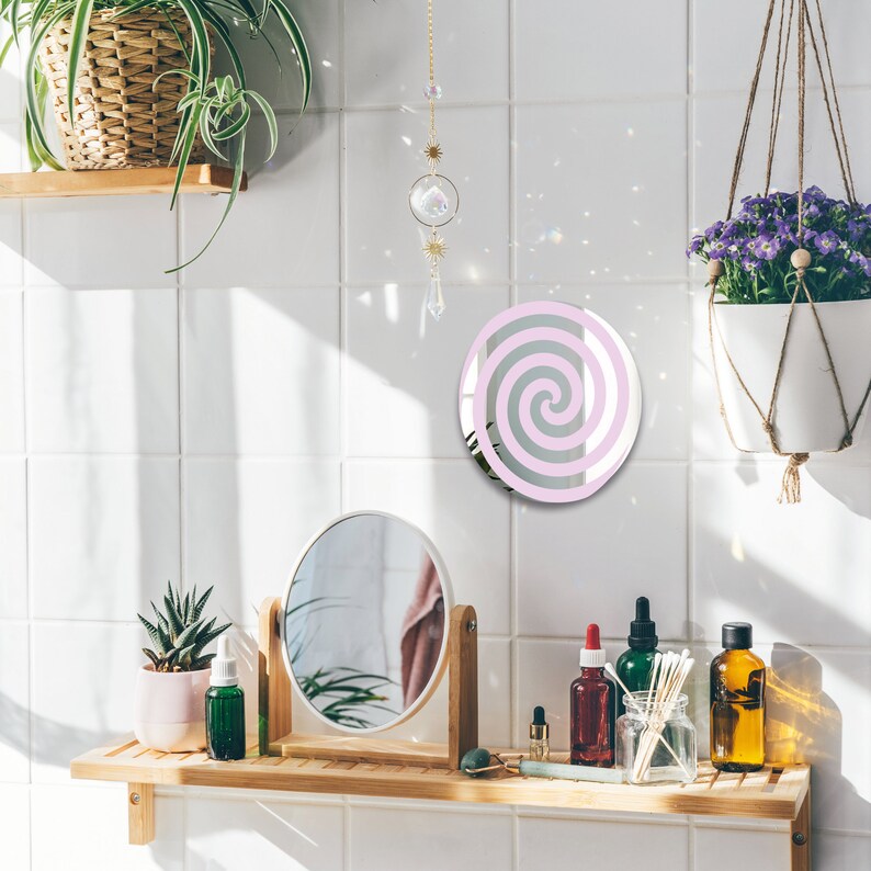 May include: A pink and white spiral design mirror with a white frame, hanging on a white tiled wall in a bathroom setting. The mirror is surrounded by a decorative crystal chain and a hanging plant.