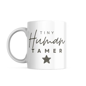 May include: White ceramic mug with a black and gray design. The mug says "Tiny Human Tamer" with a star below the text.