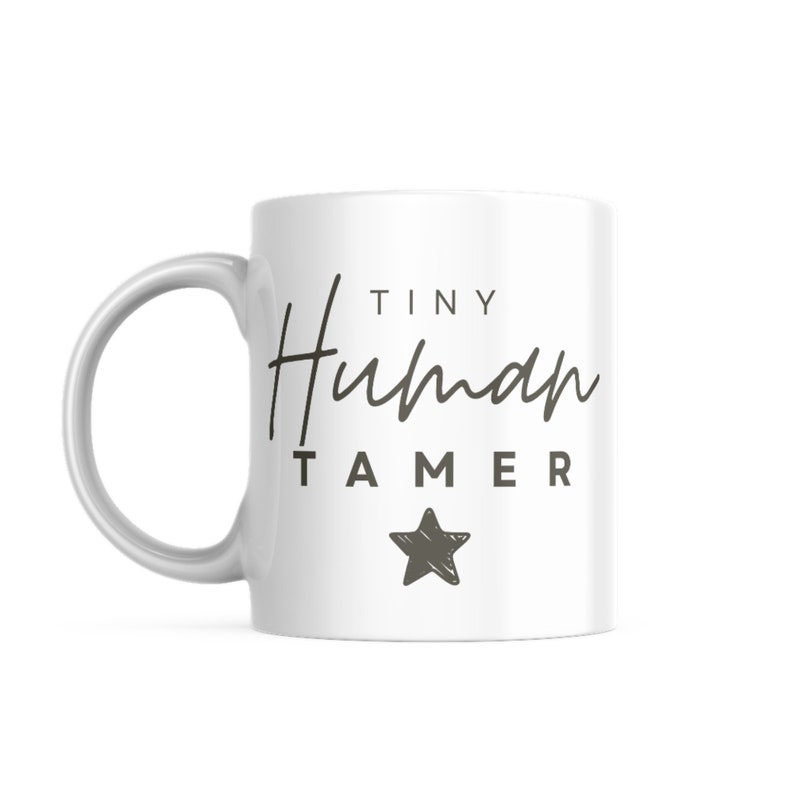 May include: White ceramic mug with a black and gray design. The mug says "Tiny Human Tamer" with a star below the text.