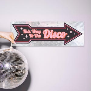 Pu&ograve; includere: Un cartello in stile retr&ograve; con la scritta "This Way To The Disco" in rosa e nero, con una freccia che punta verso destra. Il cartello &egrave; posto su uno sfondo argentato e specchiato. Una palla da discoteca &egrave; appesa sotto.