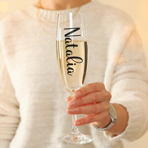 May include: A champagne flute filled with a bubbly, light-colored beverage. The glass has the name "Natalia" in a black script font. The flute is held by a person wearing a white sweater.