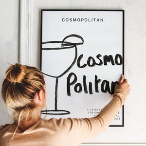 May include: A black and white illustration of a cocktail glass with the text "COSMOPOLITAN" and "COSMO Politan" written in a stylized font. The text below the illustration includes the ingredients for the cocktail: 1 1/2 oz vodka, 1 oz cranberry juice, 1/2 oz lime juice, and 1/4 oz Cointreau.