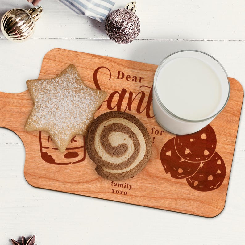May include: A wooden serving board with a handle, featuring the text "Dear Santa" and "family xoxo". It holds a star-shaped cookie, a swirl cookie, a glass of milk, and a cookie graphic. The board is light brown.
