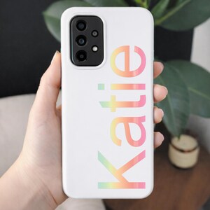 May include: White phone case with the name "Katie" in a gradient of pastel colors. The case has a camera cutout and is held in a hand.