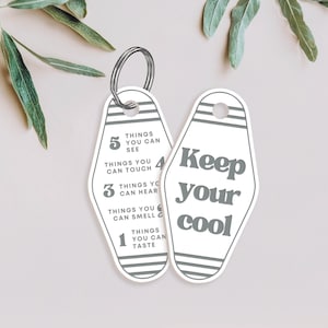 K&ouml;nnte beinhalten: Wei&szlig;er Motel-Schl&uuml;sselanh&auml;nger mit schwarzen und grauen Streifen. Der Anh&auml;nger hat auf der einen Seite den Text "Keep your cool" und auf der anderen Seite eine Liste von Dingen, die man sehen, ber&uuml;hren, h&ouml;ren, riechen und schmecken kann.