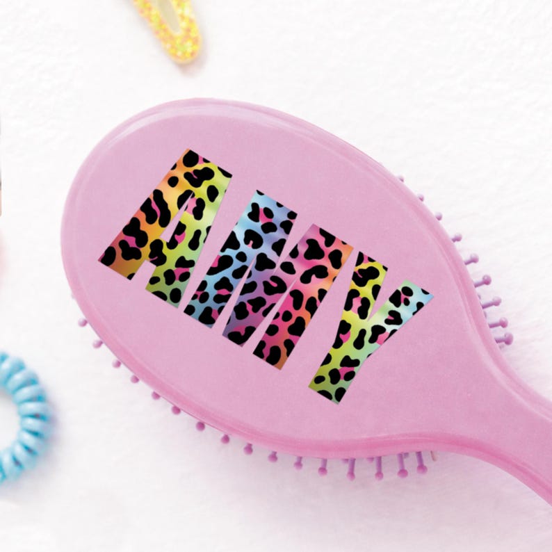 May include: A pink oval hairbrush with the name "AMY" in colorful leopard print lettering. The brush has soft bristles and is surrounded by other hair accessories.