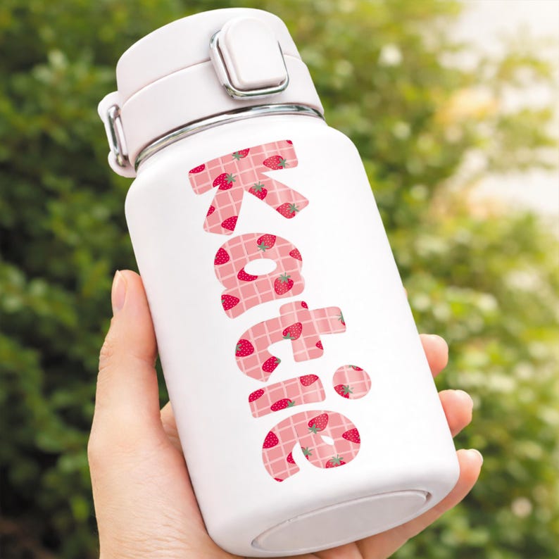 May include: White insulated water bottle with a pink lid and a silver clasp. The name "Katie" is printed in large pink letters filled with a strawberry pattern. The bottle is held against a blurred green background.