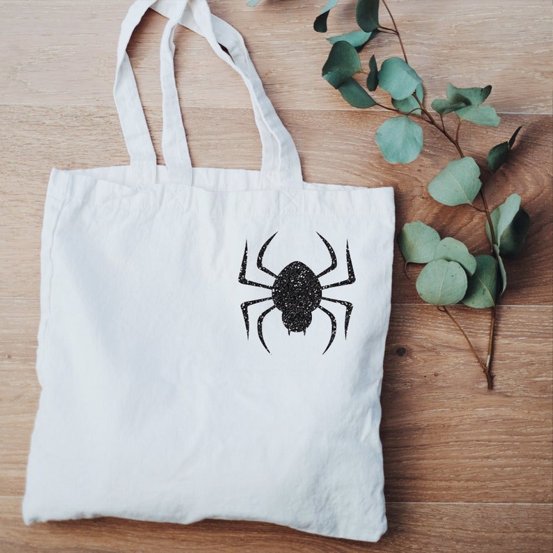 May include: A white tote bag with a black glitter spider design. The bag has two handles and is made of a soft, natural fabric.