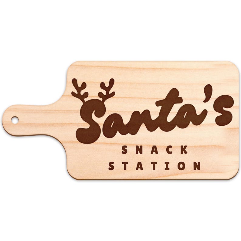 Op de afbeelding: Houten snijplank met een handvat en de tekst "Santa's Snack Station" in bruine letters. Het woord "Santa's" heeft een rendiergeweiontwerp erboven. De plank is lichtbruin met een houtnerf.