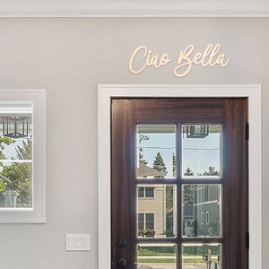 May include: A wooden sign with the text "Ciao Bella" in cursive script, mounted on a light grey wall above a brown wooden door with glass panels.