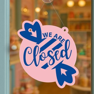 May include: A pink, circular sign with the words "WE ARE Closed" in blue script. The sign has blue decorative elements and is hanging from a string. The sign is displayed on a glass door.