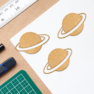 May include: Three wooden cutouts of planets with rings, each with a brown color and a white ring. The planets are on a white surface.