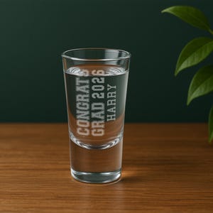 May include: A clear shot glass filled with liquid, featuring the text "CONGRATS GRAD 2026 HARRY" in a collegiate font. The glass is set on a wooden surface against a dark green background.