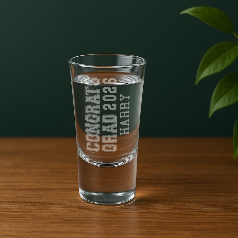 May include: A clear shot glass filled with liquid, featuring the text "CONGRATS GRAD 2026 HARRY" in a collegiate font. The glass is set on a wooden surface against a dark green background.