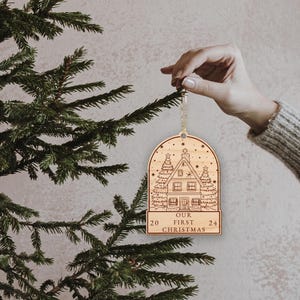 May include: Wooden Christmas ornament shaped like a snow globe with a house and trees inside. The ornament is inscribed with "Our First Christmas 20 24".