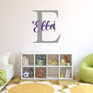 May include: A gray letter E with the name "Ella" written in purple cursive on top of it. The letter is on a white wall in a room with a white bookshelf, a green armchair, and a rug.