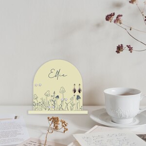 May include: A yellow wooden earring holder with a mushroom and flower design. The holder has the name "Ellie" written on it.