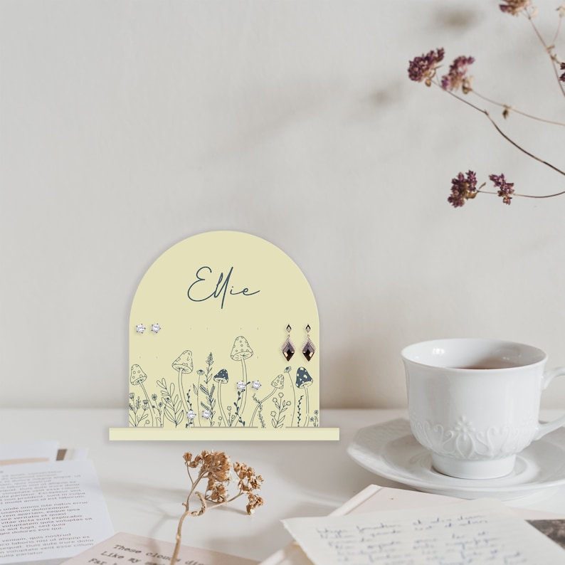 May include: A yellow wooden earring holder with a mushroom and flower design. The holder has the name "Ellie" written on it.