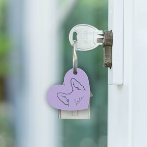 Op de afbeelding: Een paarse hartvormige sleutelhanger met een hondensilhouet en de naam "Lola" erop gegraveerd. De sleutelhanger is bevestigd aan een zilveren sleutelring en een sleutel.